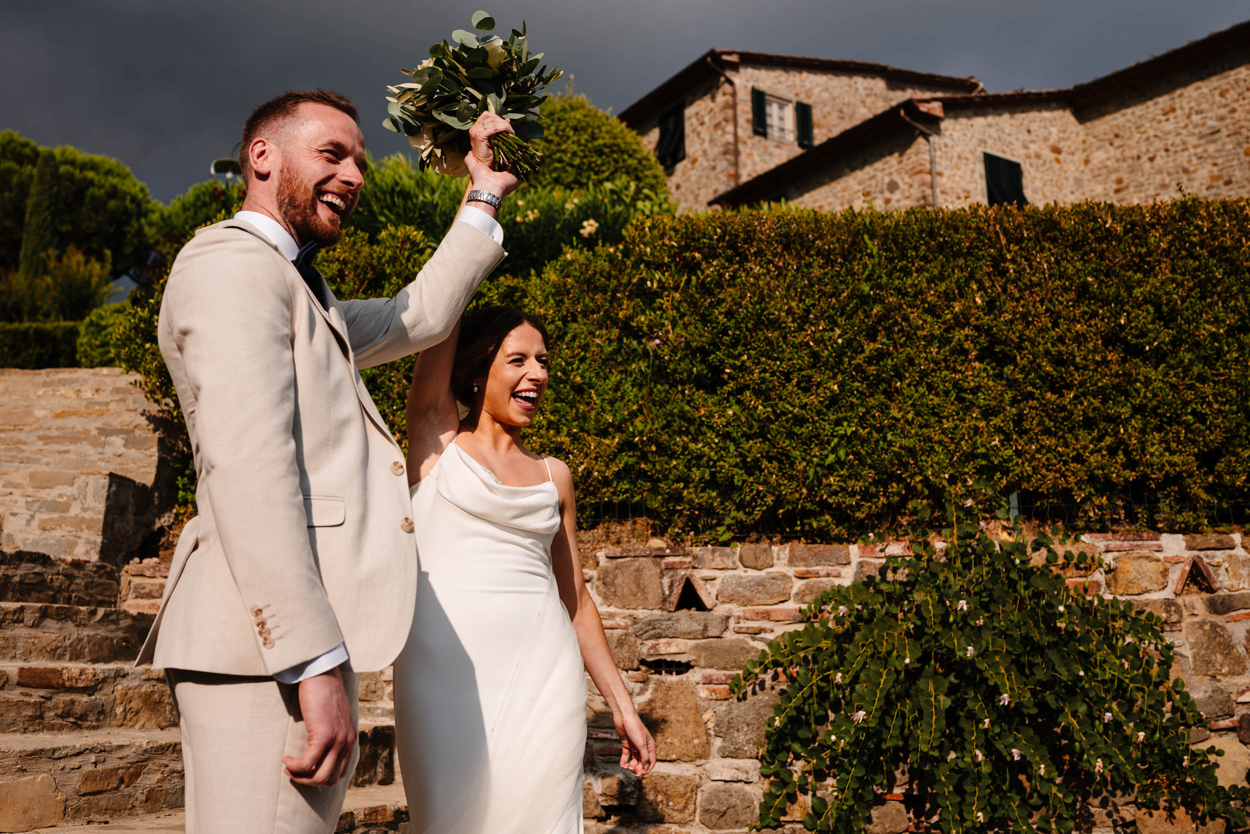 Couple laughing and raising the bouquet with the stone villa in the background.