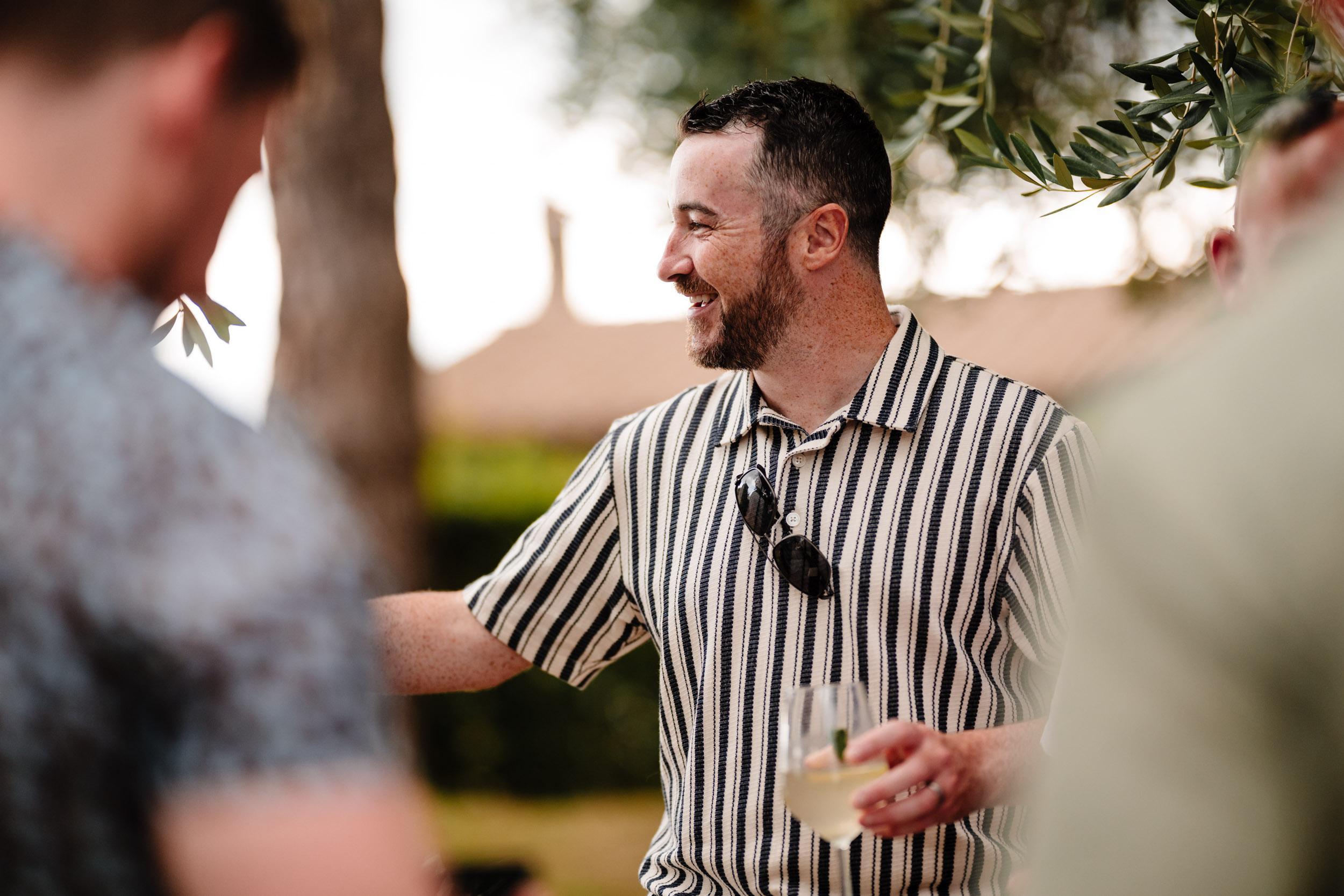 Wedding guest smiling and holding a drink under the olive trees.
