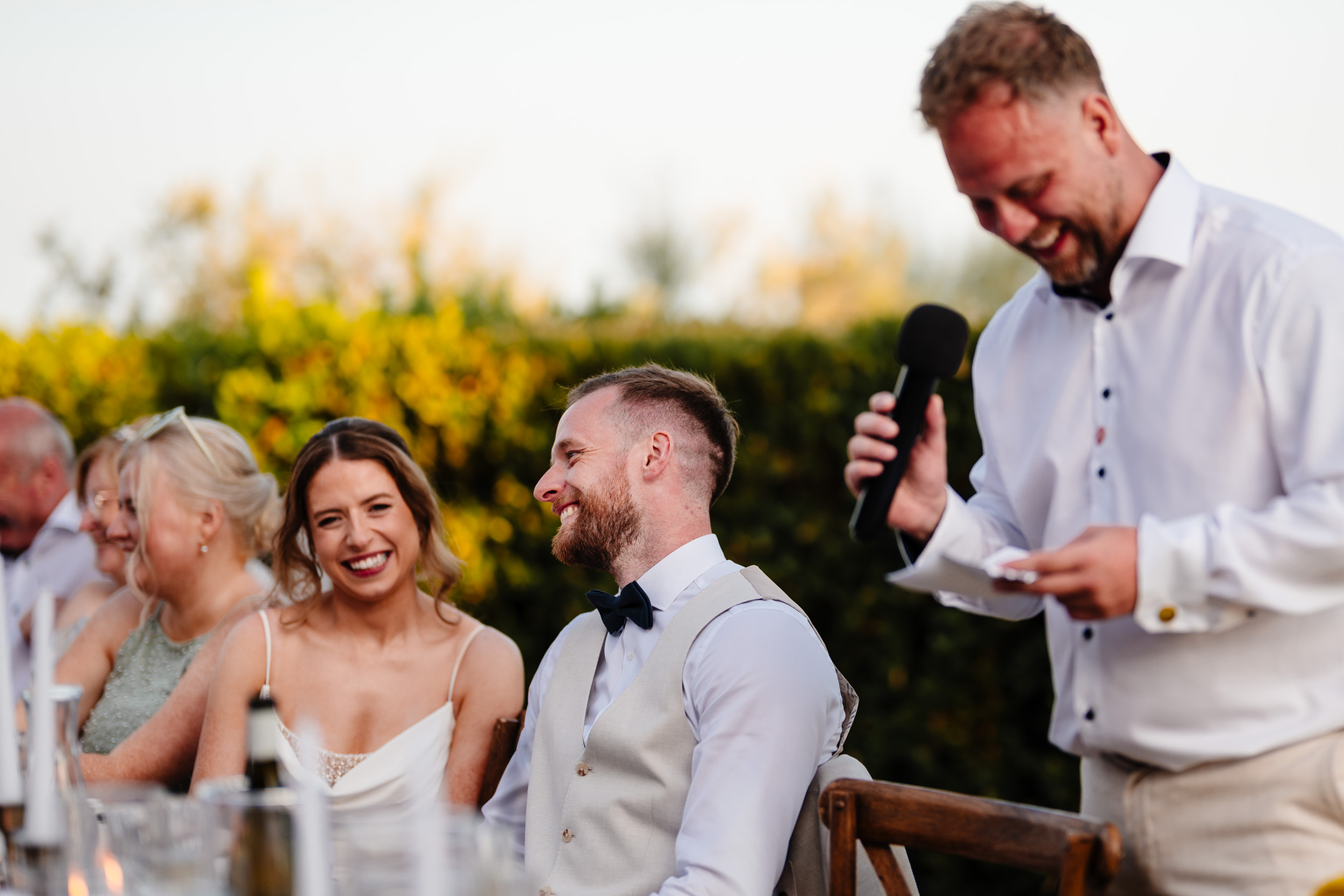 Bride and groom smiling as a guest gives a speech at the outdoor dinner table.