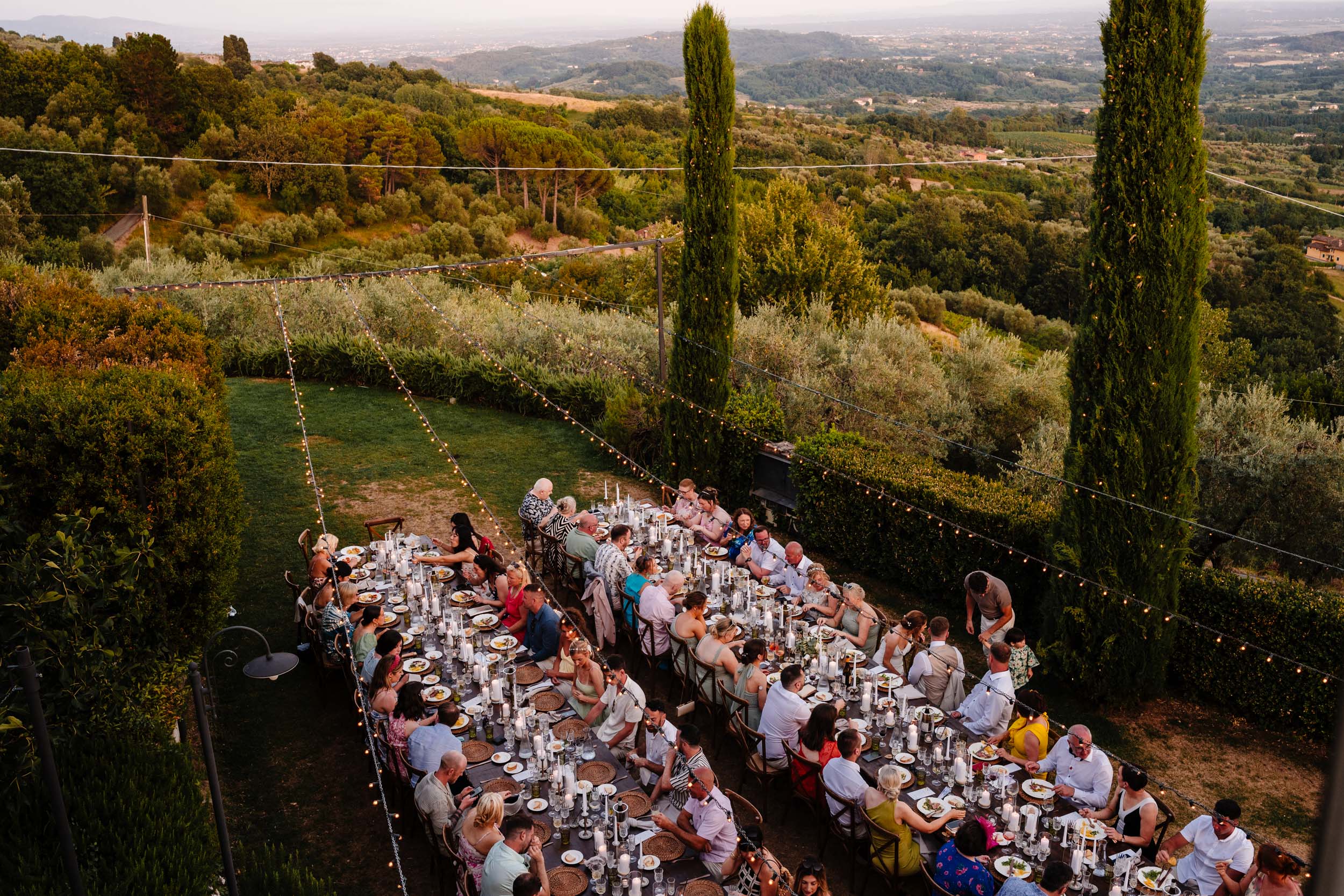 Long outdoor dinner table surrounded by guests with Tuscan hills in the background.