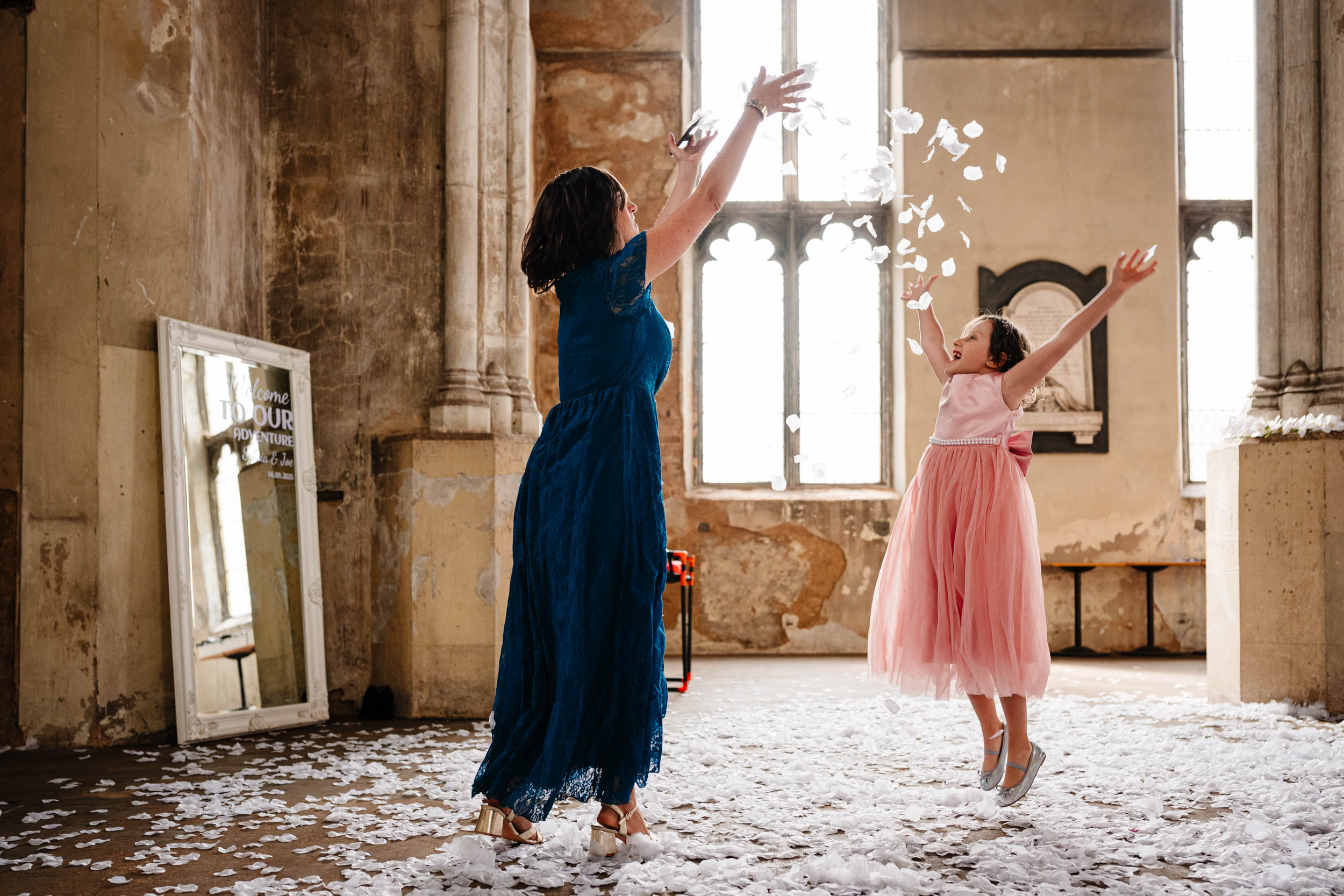 Mother and young daughter tossing flower petals into the air outside, playful candid wedding moment.