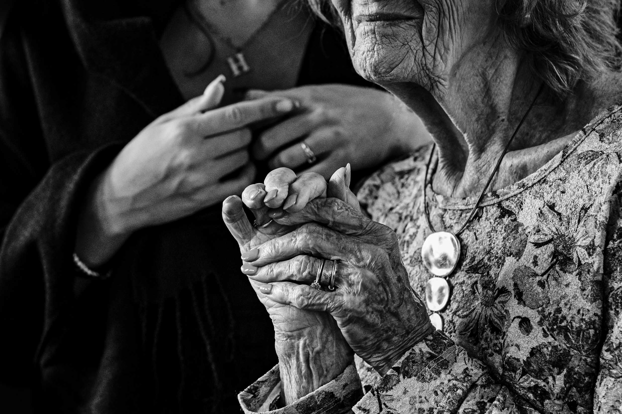Close-up detail of an older woman’s wrinkled hands and face, with a younger woman’s hands blurred in the background.