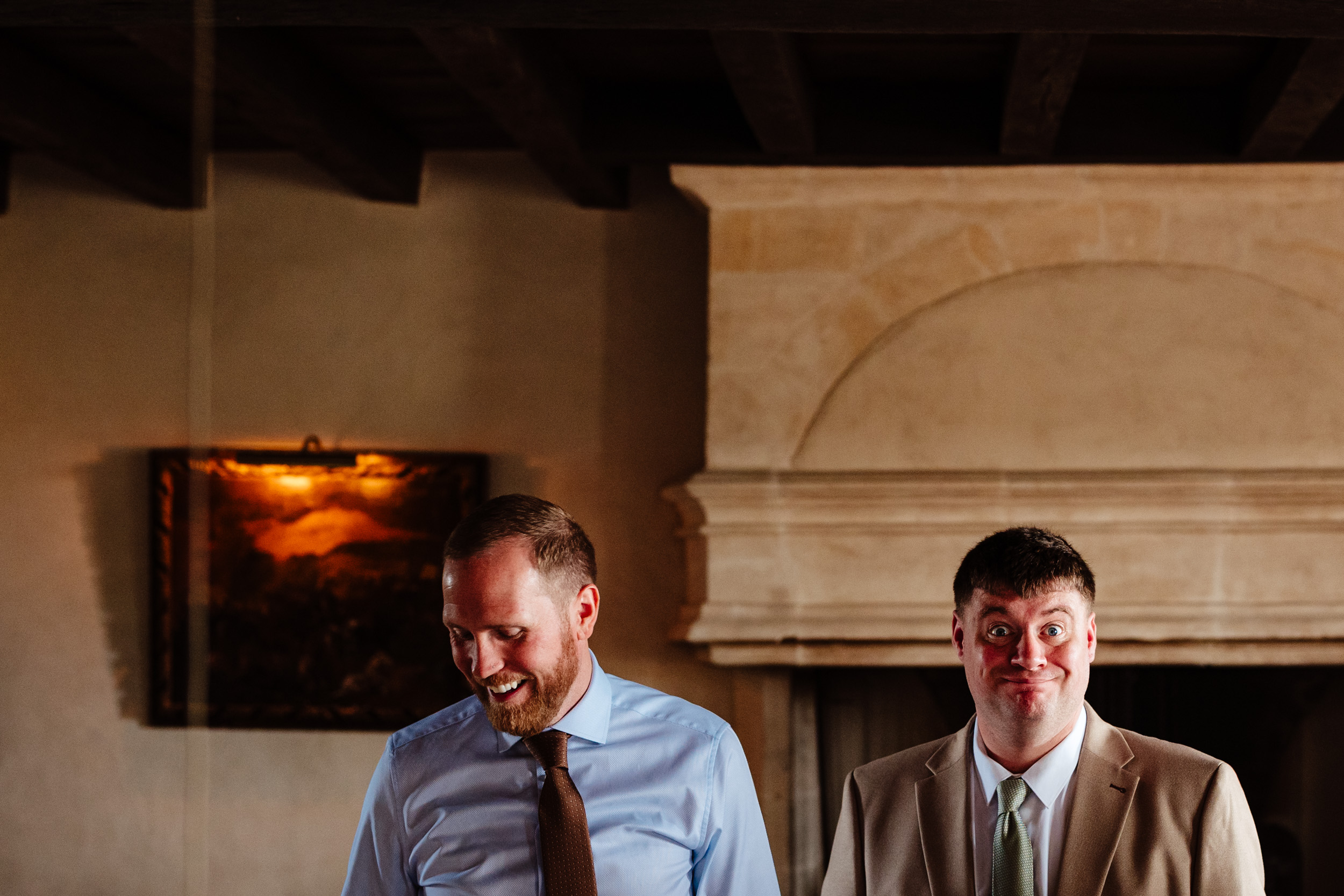 Groom looking down while his cousin pulls a funny face during relaxed wedding morning prep