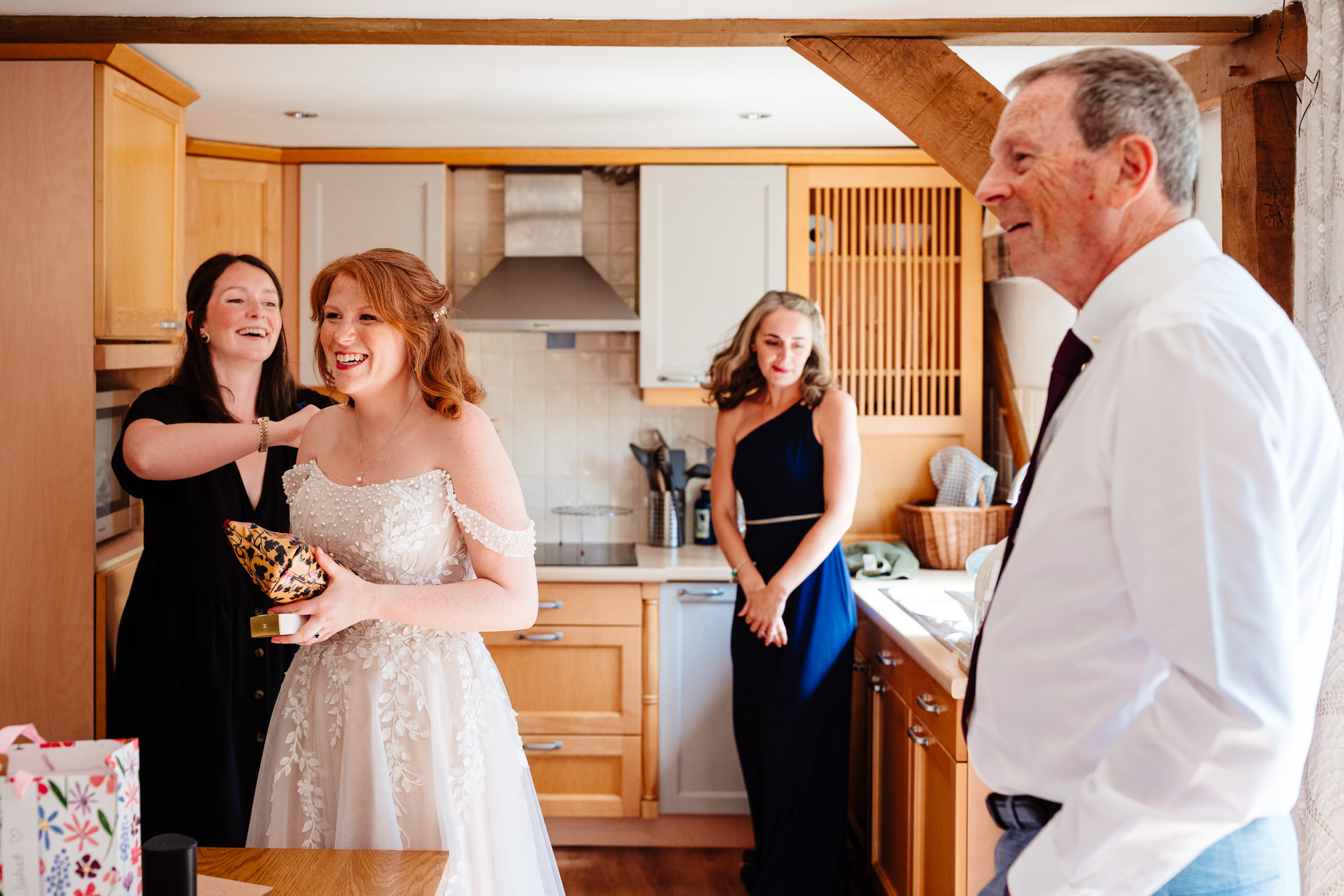 Bride in her wedding dress as a makeup artist makes final adjustments with her dad and a bridesmaid nearby