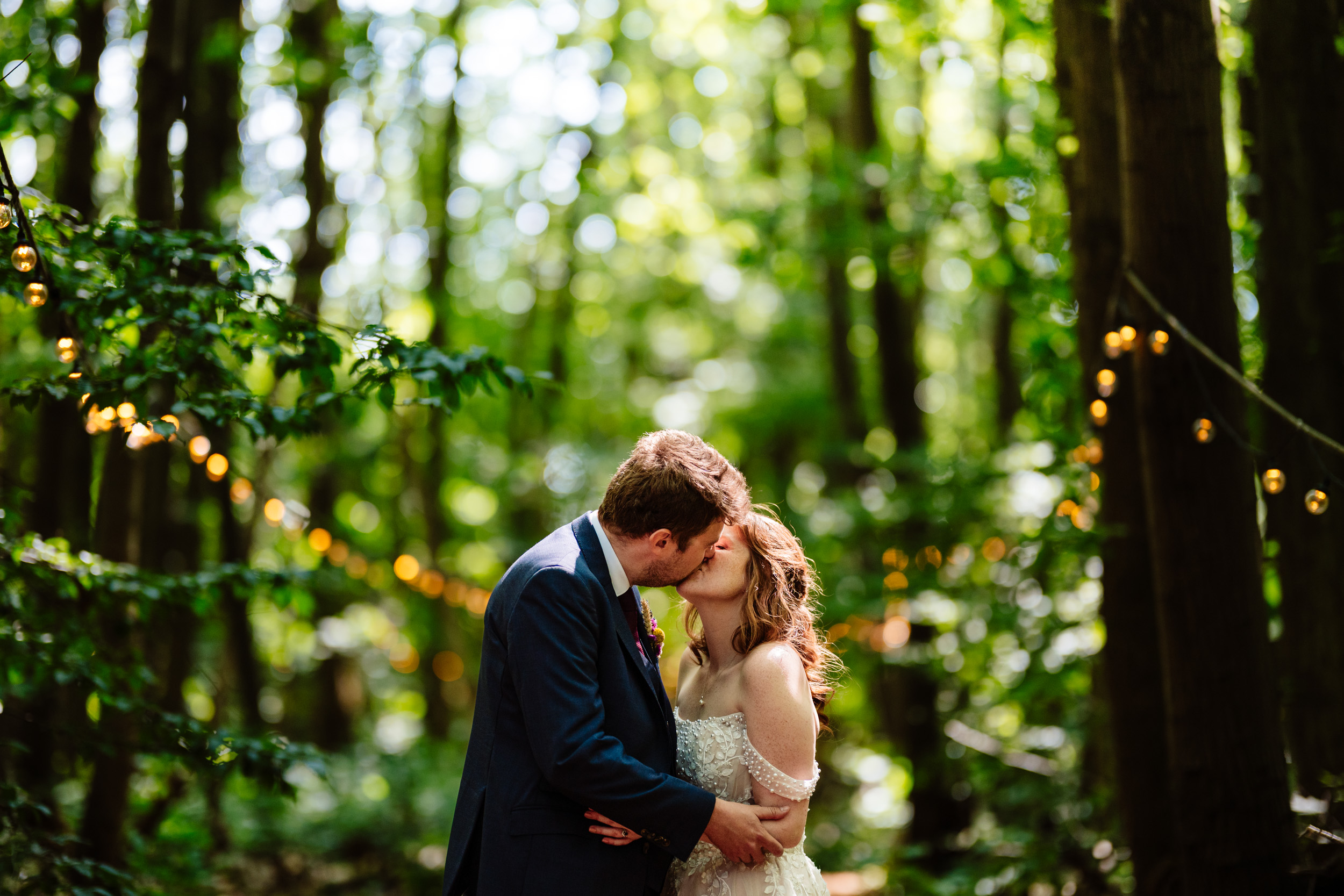 Bride and groom sharing a kiss during couple portraits on the festoon-lit path at The Dreys