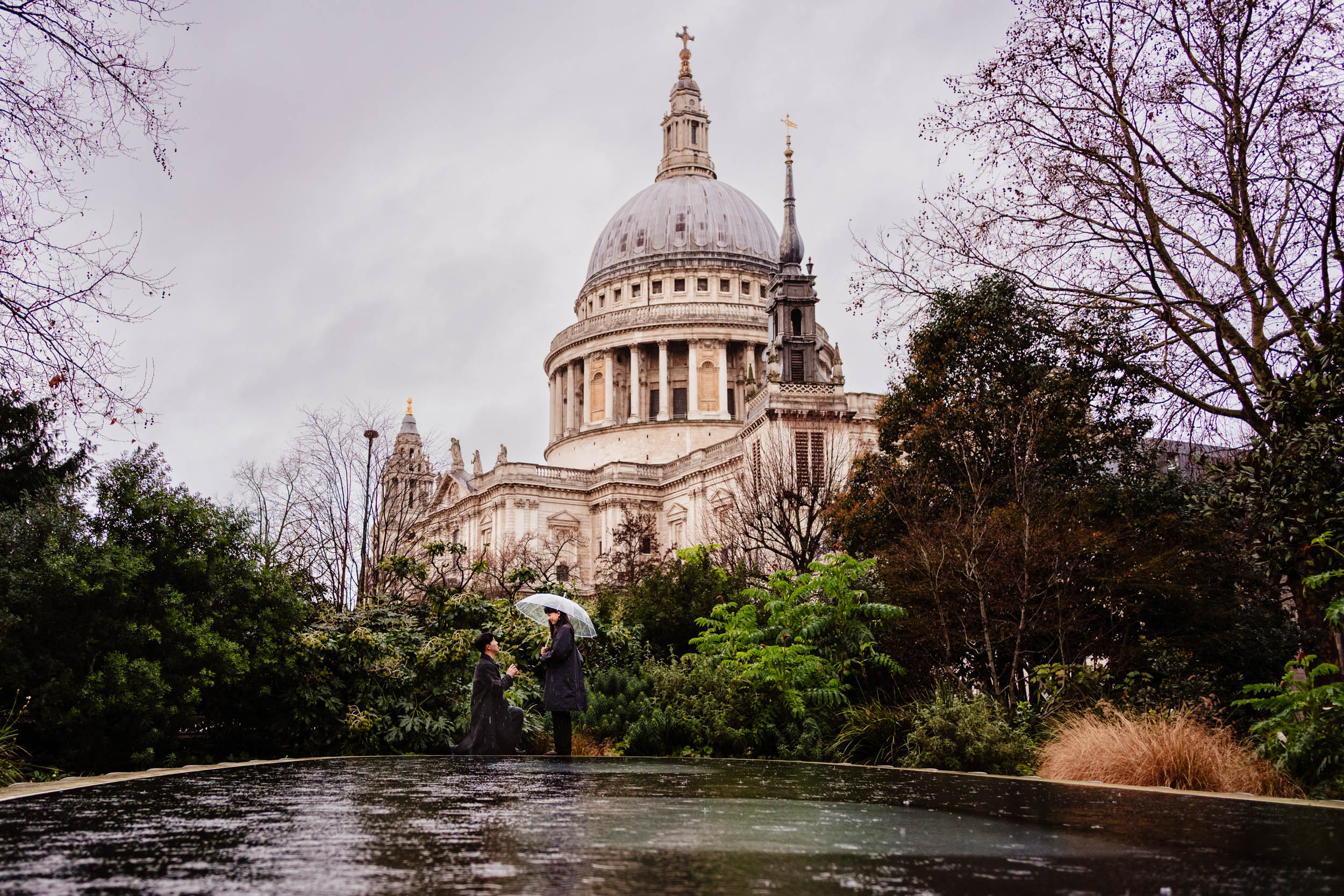 Couple during their surprise proposal at St Paul’s Cathedral with the Reflection Garden pool in the foreground.