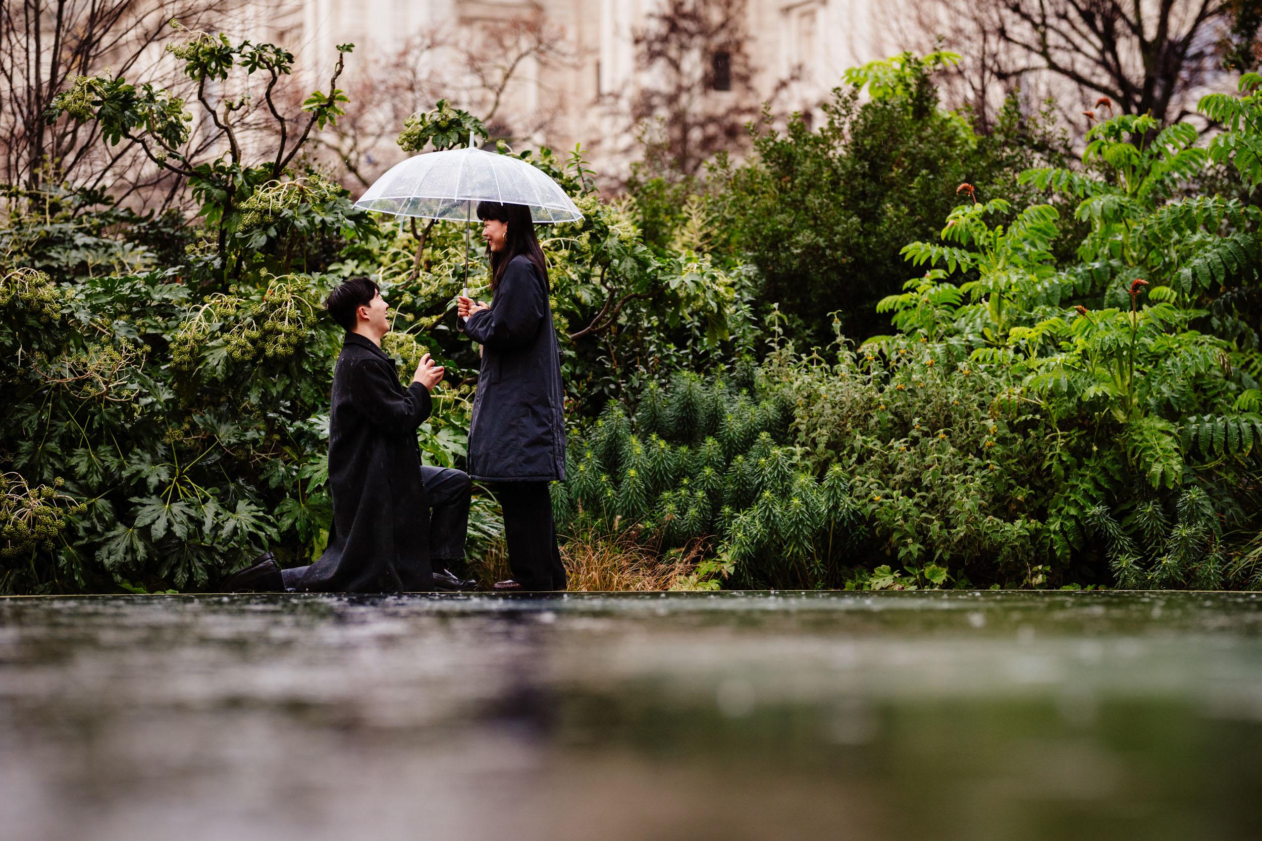 Close-up of a surprise proposal at St Paul’s Cathedral in the rain, with a clear umbrella as he kneels.