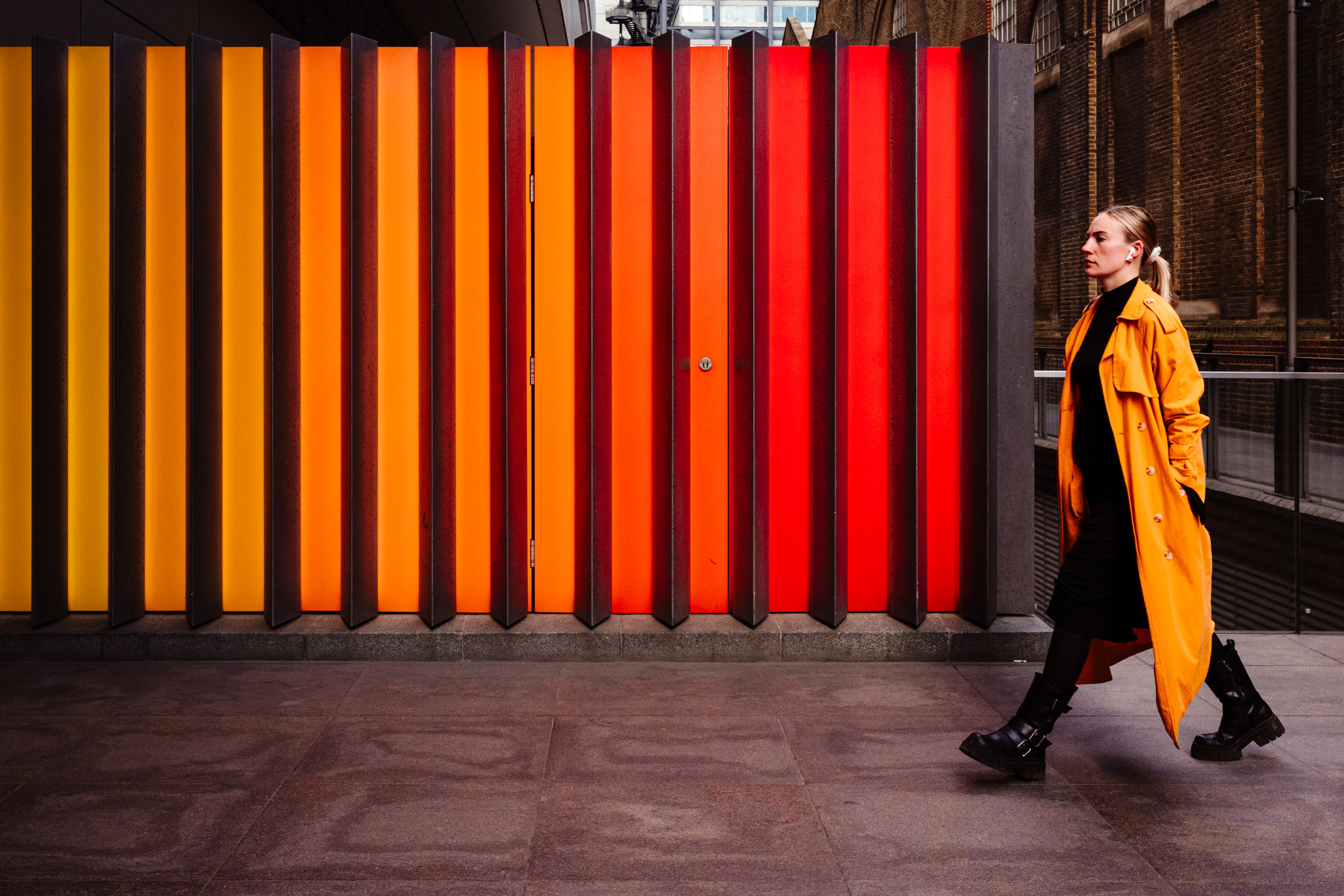 Woman in an orange coat walks in front of a similar coloured wall.