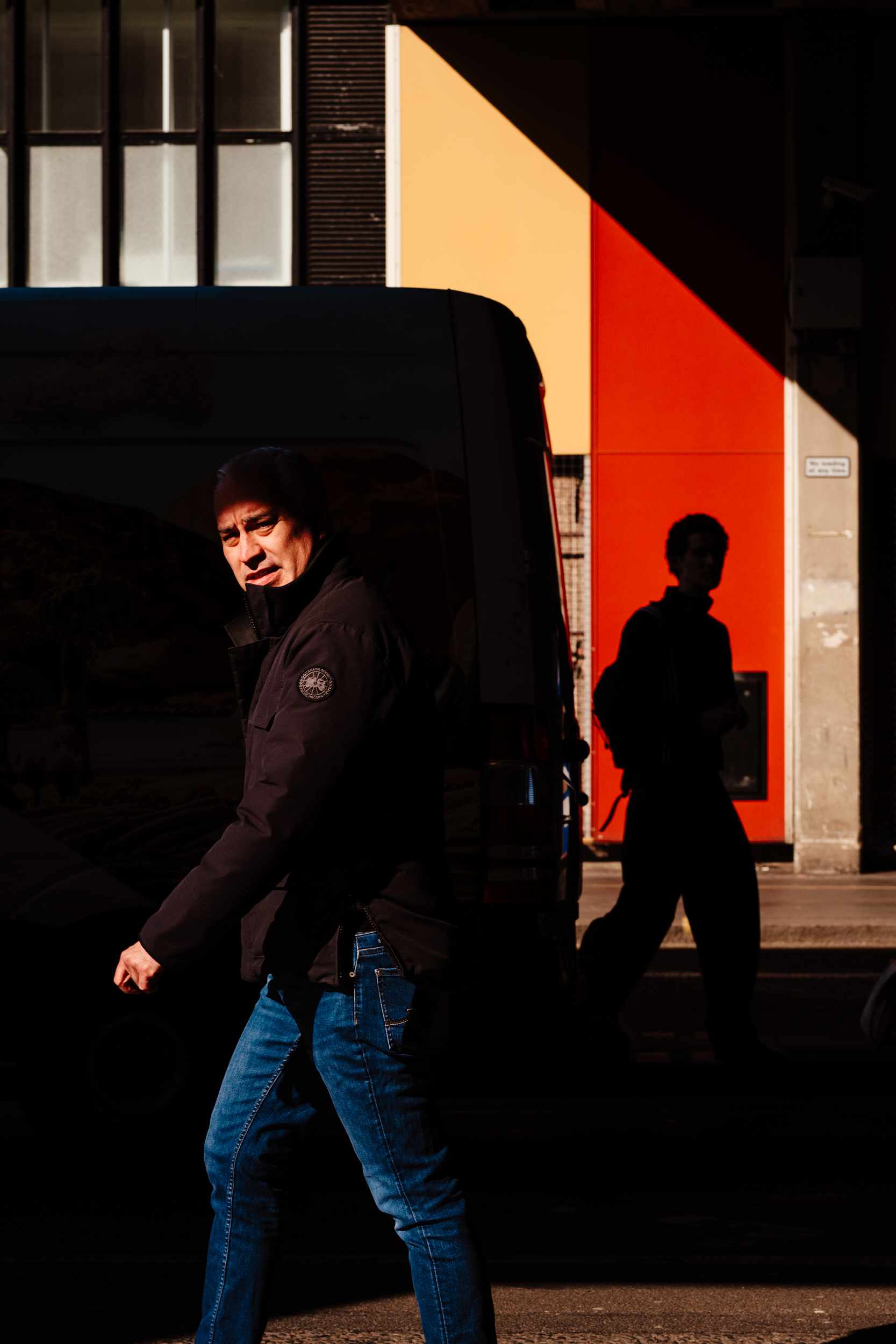 A man looks at the camera while somebody is walking behind him in the opposite direction.